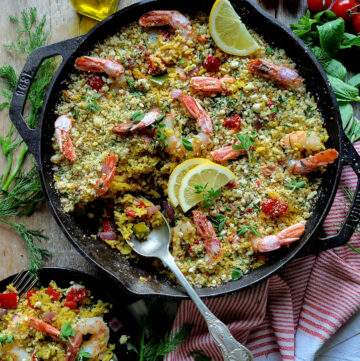 A skillet is filled with Mediterranean Shrimp and Rice Bake, with a portion spooned into a plate nearby. A bottle of olive oil and fresh herbs surround the skillet.