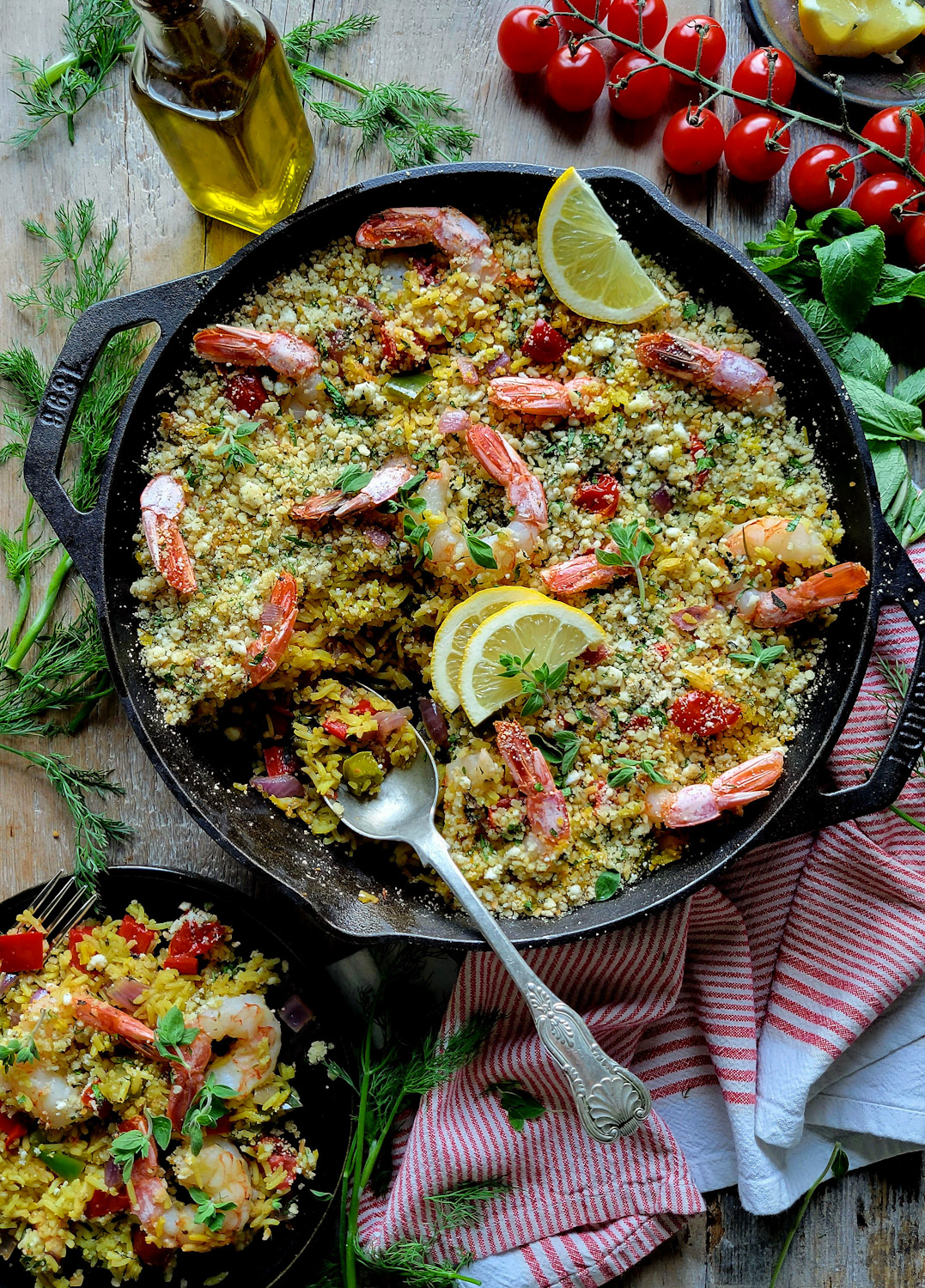 A skillet is filled with Mediterranean Shrimp and Rice Bake, with a portion spooned into a plate nearby. A bottle of olive oil and fresh herbs surround the skillet.