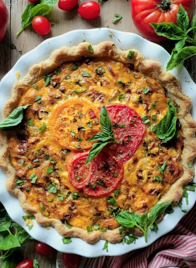 A fully baked Cheesy Tomato Pie, with fresh tomatoes and basil strewn around the pie plate.