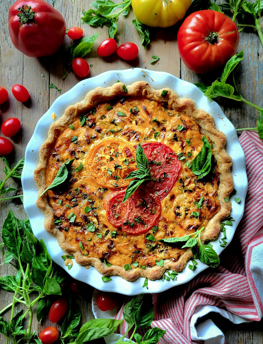 A fully baked Cheesy Tomato Pie, with fresh tomatoes and basil strewn around the pie plate.