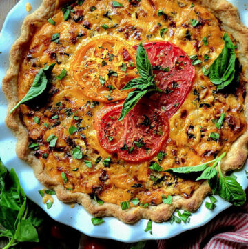 A fully baked Cheesy Tomato Pie, with fresh tomatoes and basil strewn around the pie plate.