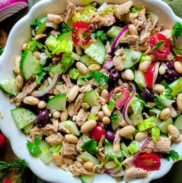 A bowl filled with Mediterranean White Bean and Tuna Salad, with olives, tomatoes, and serving utensils on the side.