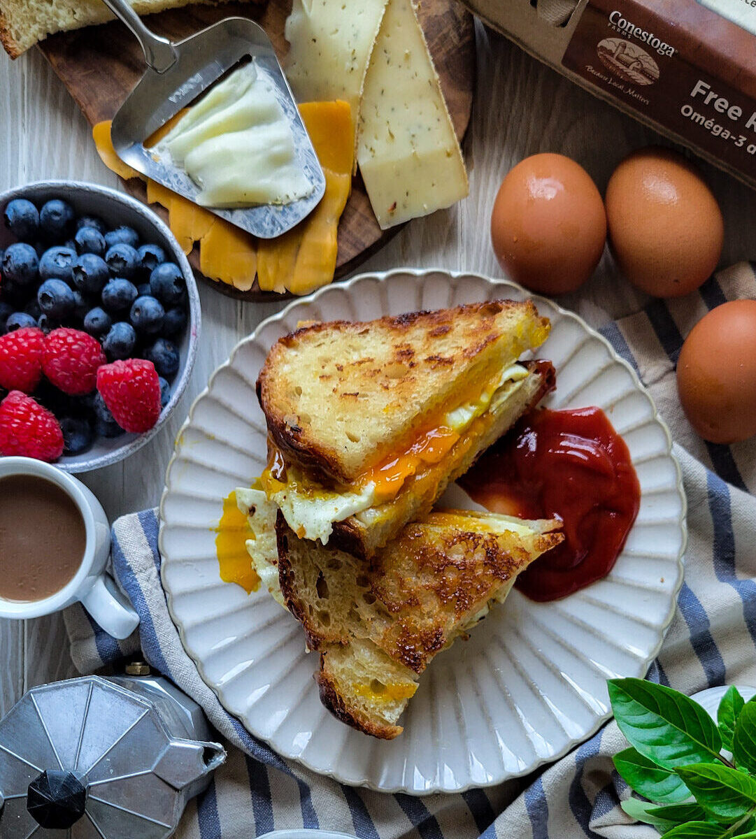 Fried Egg Grilled Cheese sandwich on a plate, surrounded by a bowl of fruit, espresso, cheese and eggs.