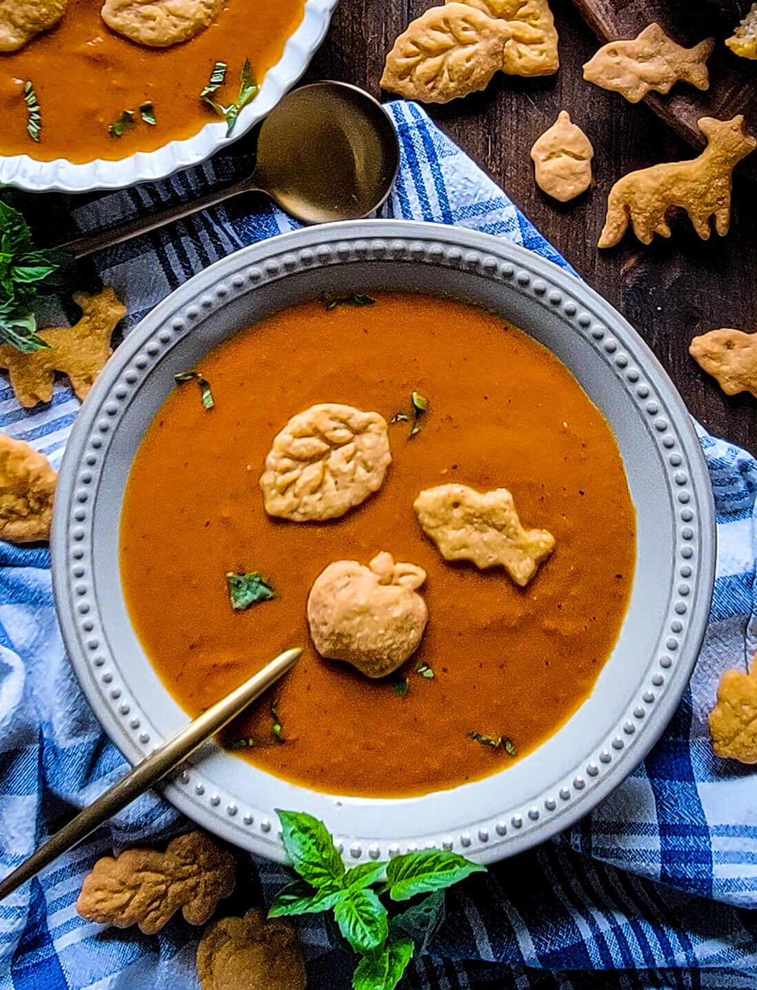 A bowl of tomato soup is topped with gluten free goldfish. More goldfish, as well as other shapes surround the bowl which is sitting on a blue and white checked cloth.