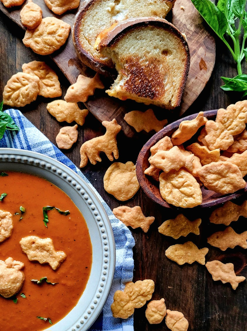 A bowl of tomato soup is surrounded by gluten free goldfish and gold leaves etc, as well as a grilled cheese sandwich.