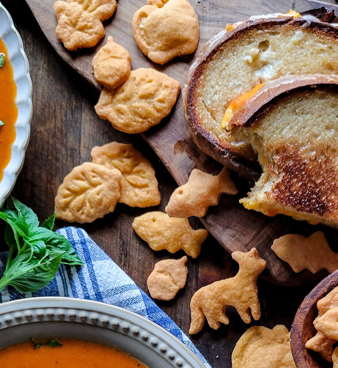 Gluten free goldfish, as well as other cheddar shapes are sitting with a grilled cheese sandwich and a bowl of tomato soup.