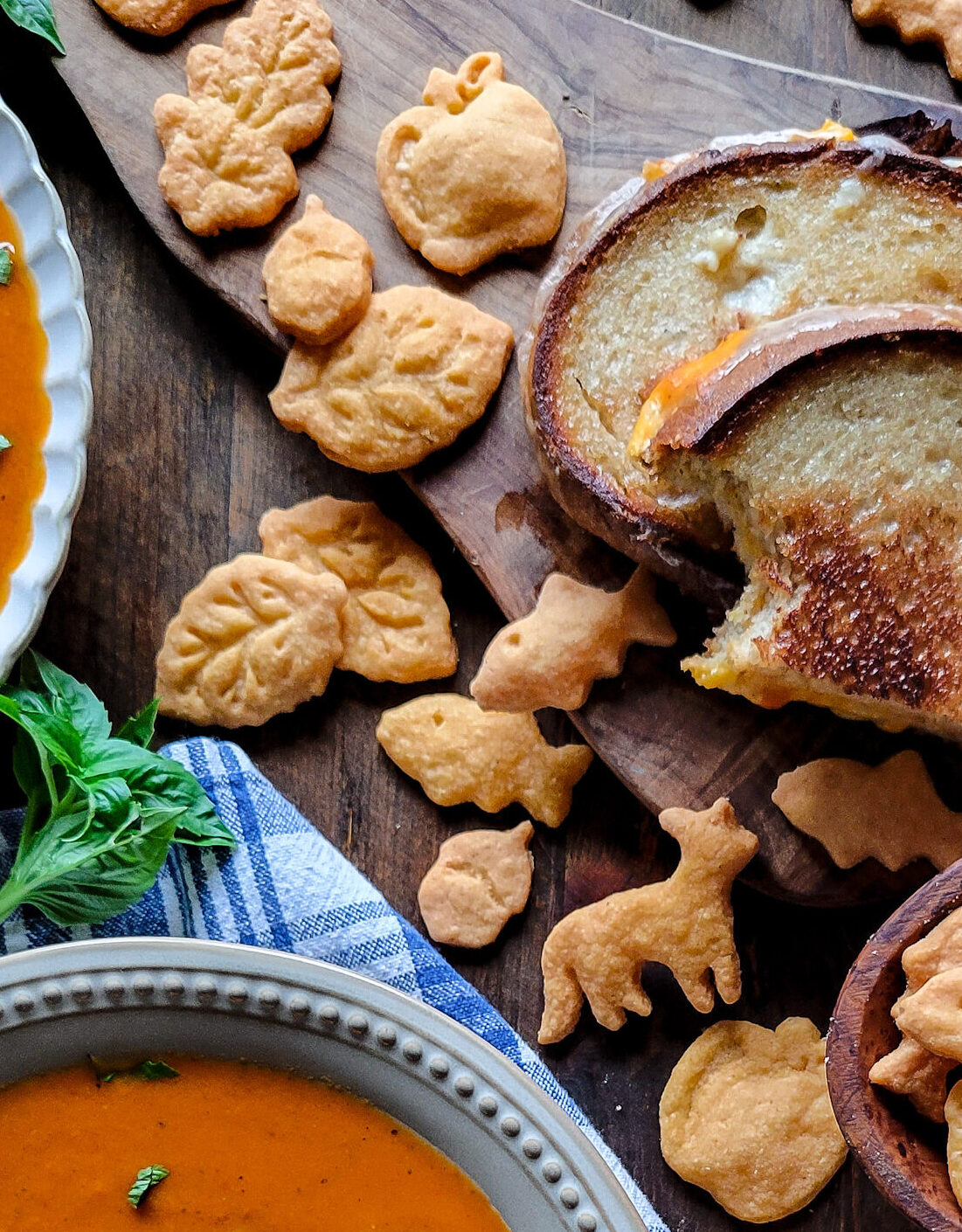 Gluten free goldfish, as well as other cheddar shapes are sitting with a grilled cheese sandwich and a bowl of tomato soup.