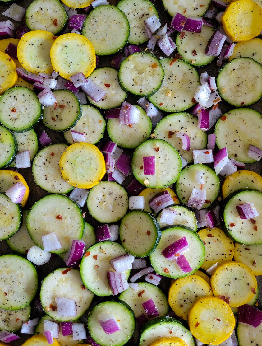 Zucchini slices and red onion spread out on a sheet pan getting ready to be baked in the oven for a Zucchini Spanakopita Quiche