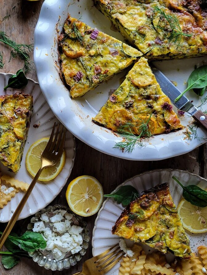 A baked Zucchini Spanakopita Quiche on the counter with lemon slices and spinach leaves strewn about. A slice has been removed and placed on a plate with lemon slices and fries.