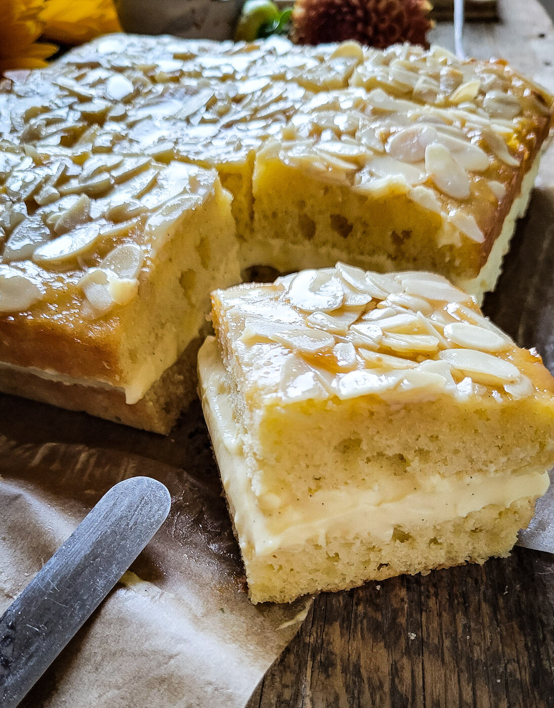 Close up of a slice of Bienenstich Torte, sitting on the cutting board, with the rest of the cake behind it.