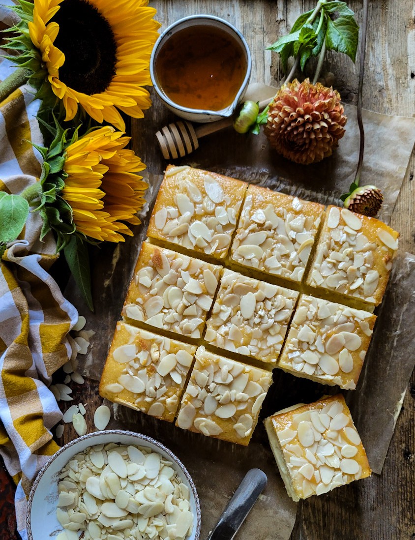 Sliced Bienenstich Torte on the cutting board, with sunflowers on the side, and a bowl of sliced almonds nearby.