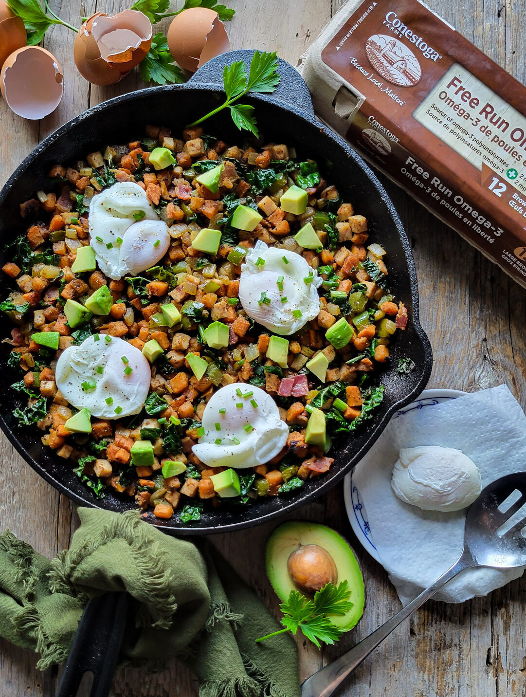 A skillet filled with Turkey Sweet Potato Hash and topped with poaches eggs. Another poached egg is on a plate, as well as avocado and and egg carton are to the side.