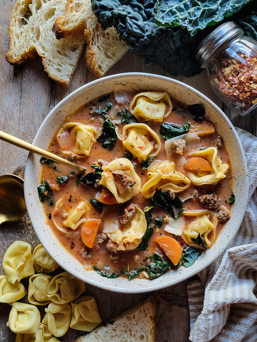 A bowl of Sausage Tortellini Soup on the table, with kale leaves, bread slices and tortellini pasta scattered about.