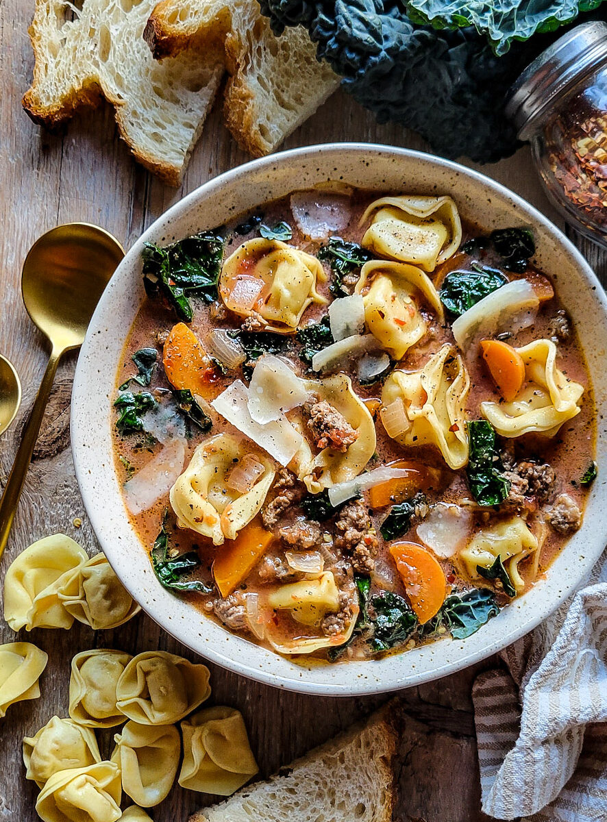 A bowl of Sausage Tortellini Soup, with slices of bread, and uncooked tortellini pasta surrounding the bowl.