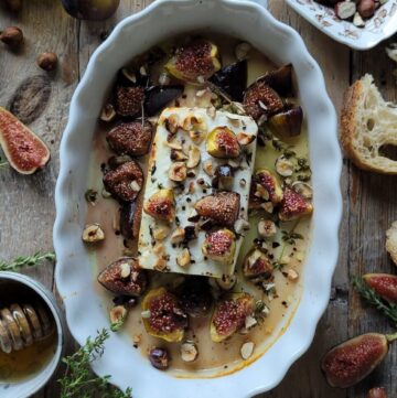 A baking dish filled with Baked Feta with Hazelnuts and Figs. Honey and fresh figs as well as herbs surround the dish.