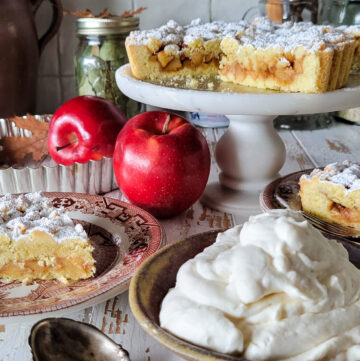 A Salted Bourbon caramel Apple Crostata is on a cake stand, with a slice on a plate and maple whipped cream in a bowl nearby.