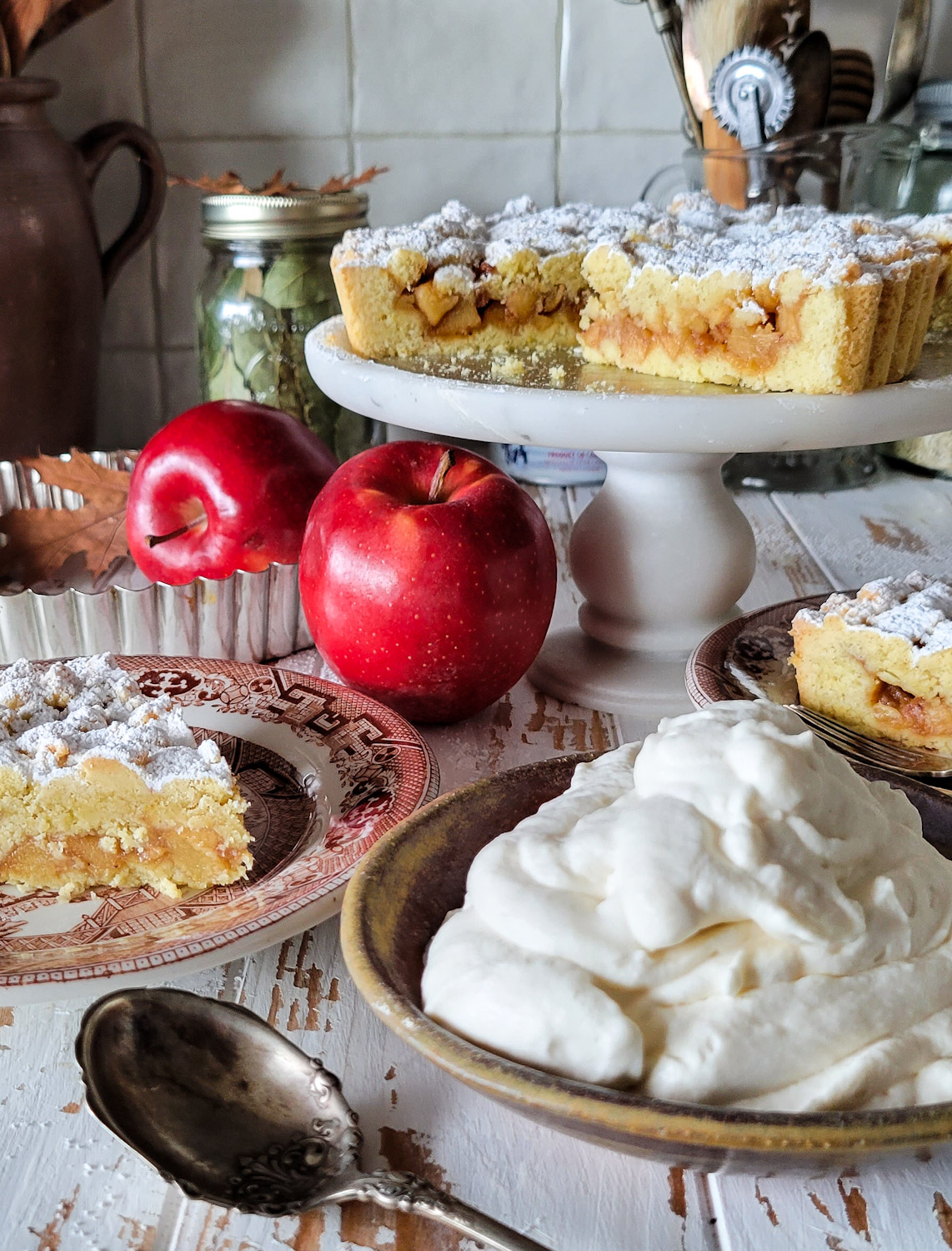 A Salted Bourbon caramel Apple Crostata is on a cake stand, with a slice on a plate and maple whipped cream in a bowl nearby.