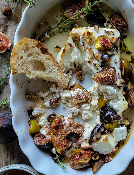 A baking dish filled with Baked Feta with Hazelnuts and Figs, and a torn piece of bread scooping up the combination. Honey and fresh figs as well as herbs surround the dish.