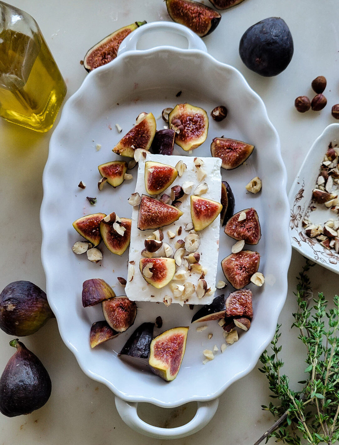 A baking dish with feta cheese, chopped hazelnuts and figs is getting ready for the oven. Olive oil, thyme and more figs surround the dish.