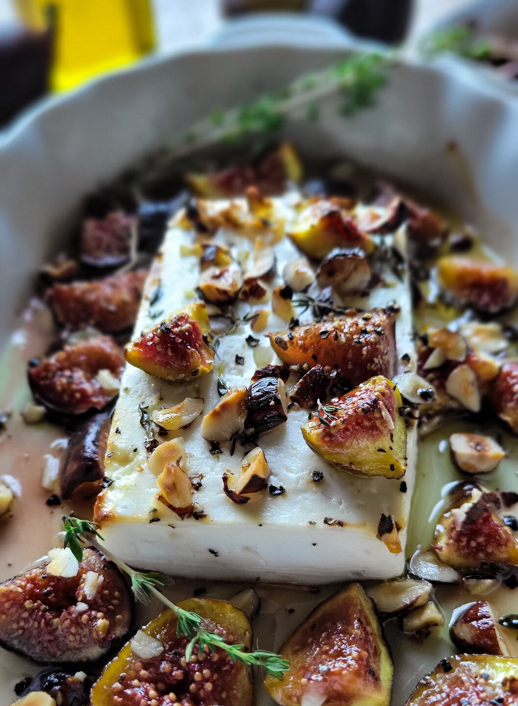 A baking dish filled with Baked Feta with Hazelnuts and Figs. Honey and fresh figs as well as herbs surround the dish.