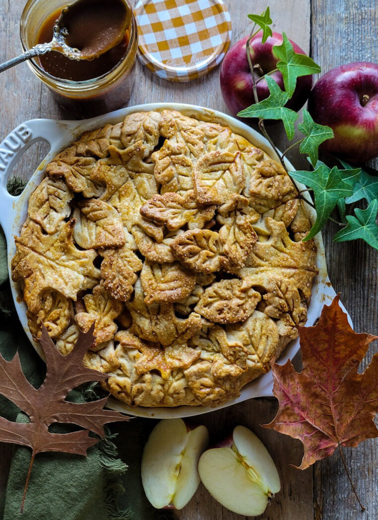 Salted Bourbon Caramel Apple Pie surrounded by leaves and apples, and a jar of caramel sauce.