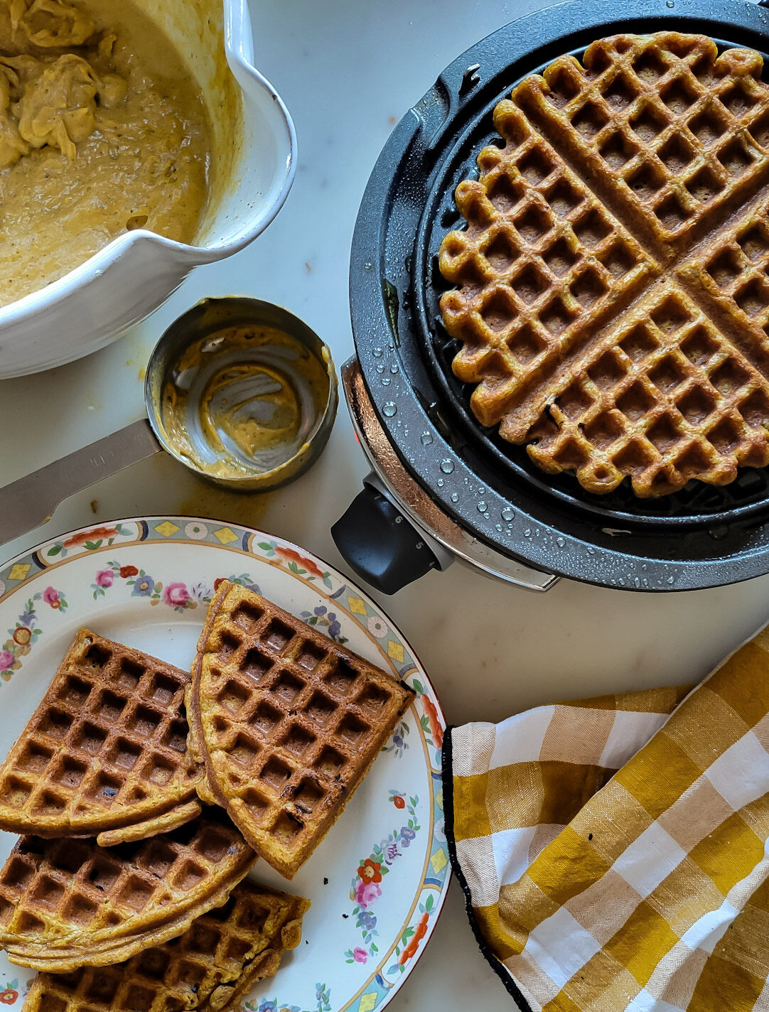 Finished Pumpkin Waffles in the waffle iron, with a plate of finished waffles on a plate, as well as the bowl of batter to the side.