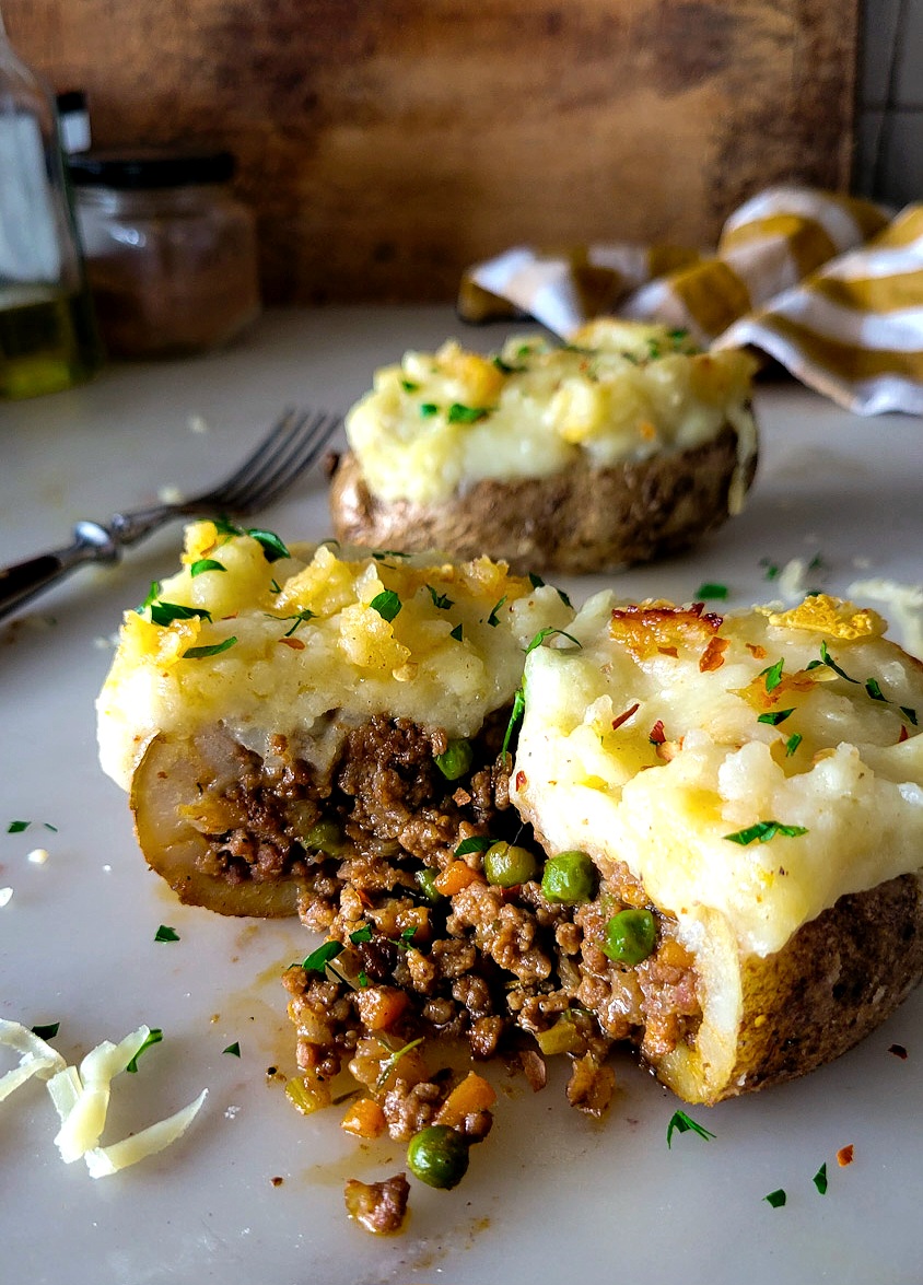 Two Shepherd's Pie Stuffed Potatoes are on the counter, one is cut open to reveal the meaty filling.