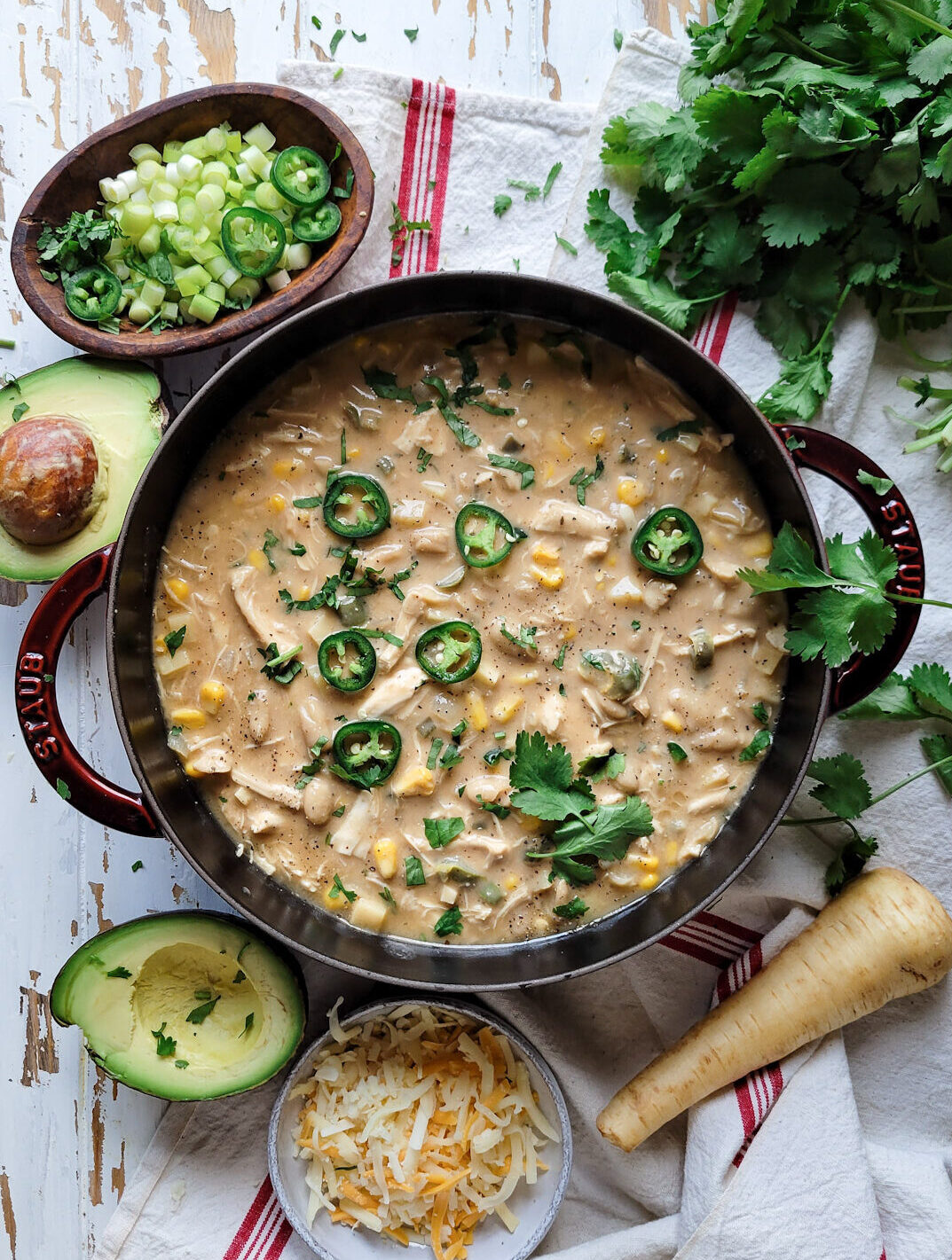 A pot of White Creole Chicken and Bean Chilli is surrounded by cilantro, chopped green onions, jalapeno pepper slices, avocado and cheese