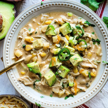 A bowl of White Creole Chicken and Bean Chilli is topped with cubed avocado, cheese and jalapeno pepper slices.