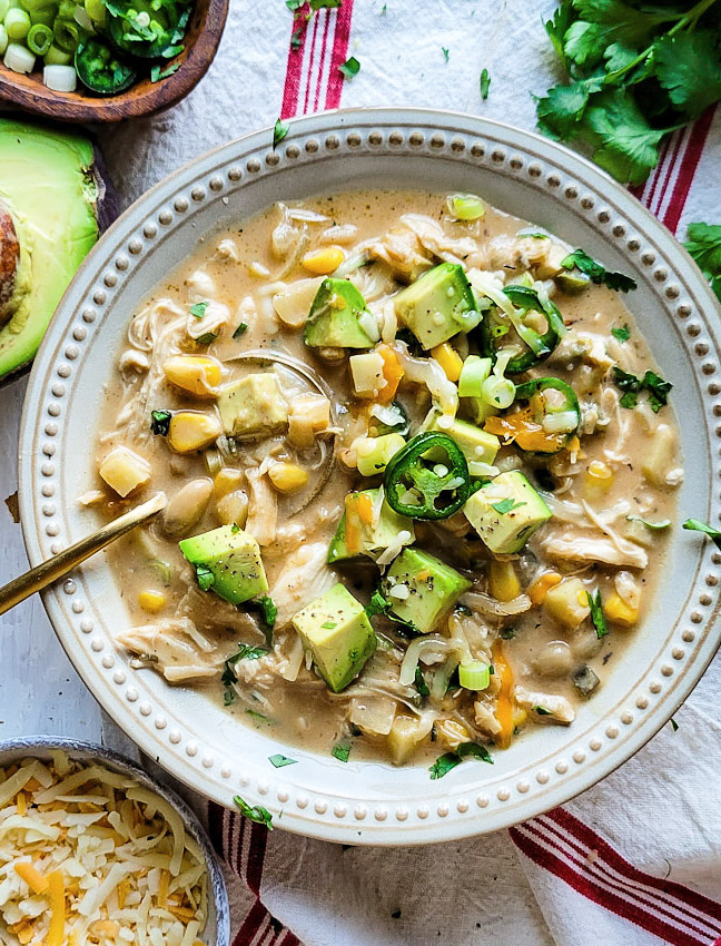 A bowl of White Creole Chicken and Bean Chilli is topped with cubed avocado, cheese and jalapeno pepper slices.