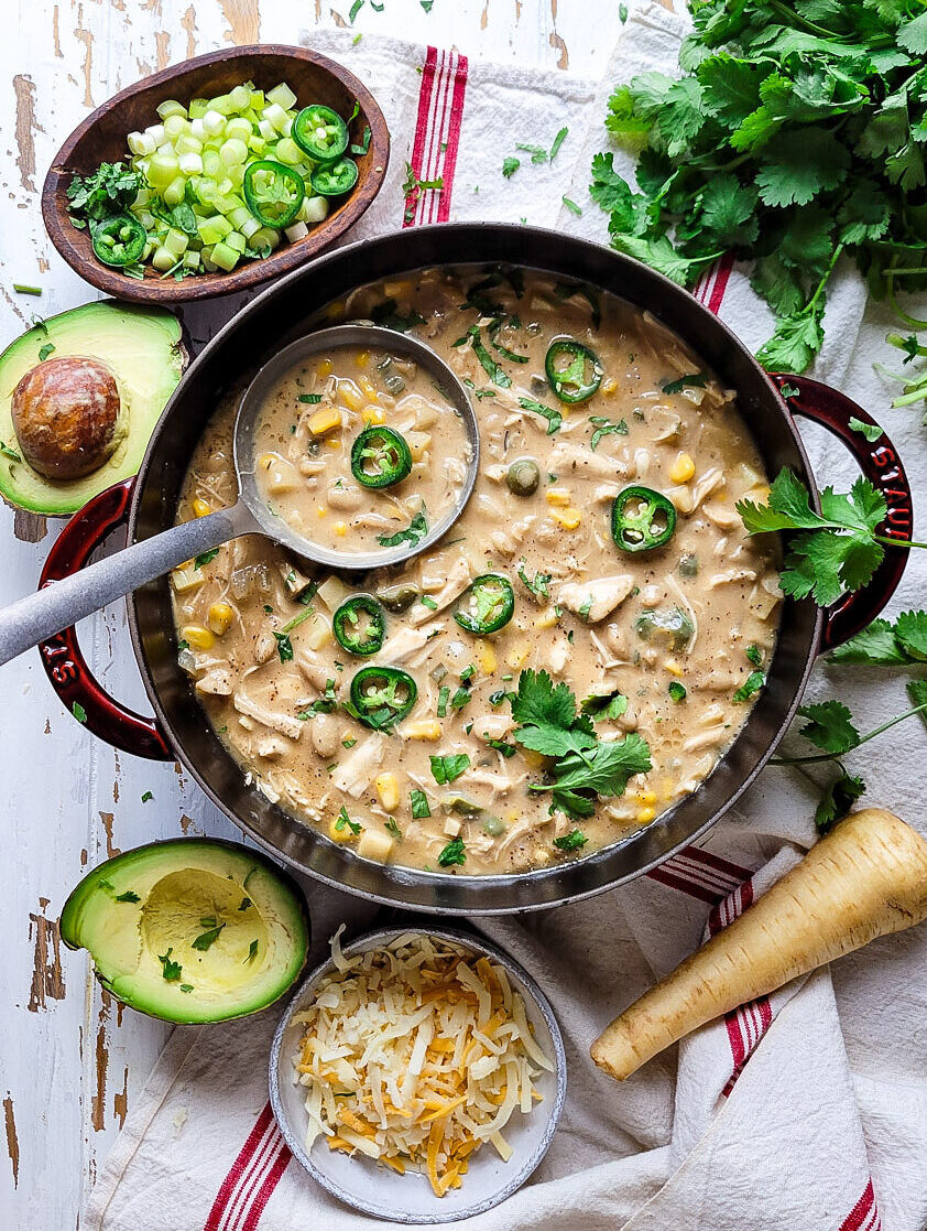 A pot of White Creole Chicken and Bean Chilli is surrounded by cilantro, chopped green onions, jalapeno pepper slices, avocado and cheese