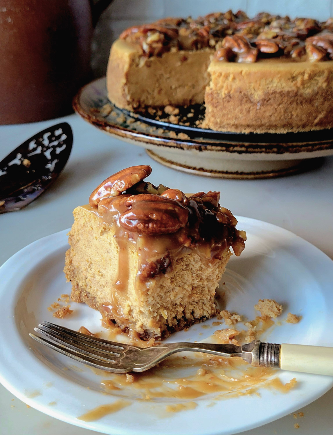 A slice of partially eaten Pecan Praline Pumpkin Cheesecake in on a plate with a fork, the rest of the cake in the background.