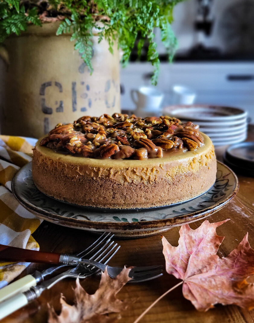 A Pecan Praline Pumpkin Cheesecake sitting on a cake stand, with serving plates and forks to the side.