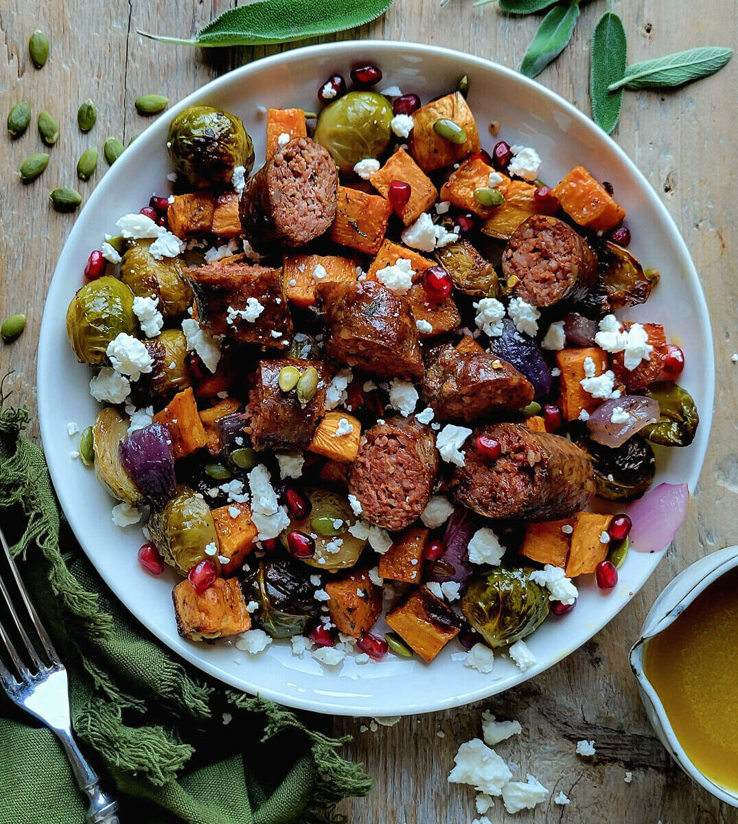 A plate filled with Roasted Autumn vegetables and Sausages, with a jar of Maple Vinaigrette to the side. Sage leaves and pumpkin seeds are scattered about.