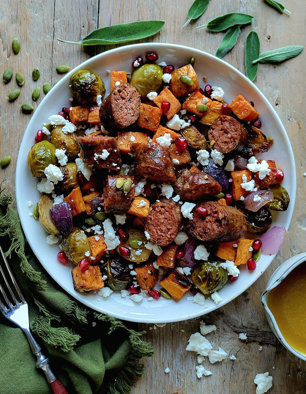 A plate filled with Roasted Autumn vegetables and Sausages, with a jar of Maple Vinaigrette to the side. Sage leaves and pumpkin seeds are scattered about.