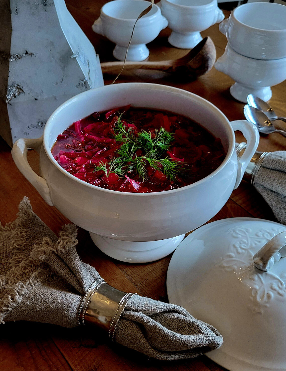 A soup tureen filled with Borscht in Short Rib Stock with soup bowls in the background.