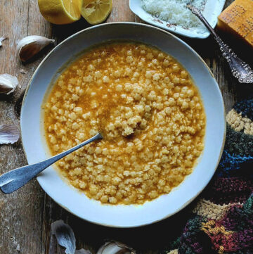 A cozy bowl of Pastina on the table with grated cheese, lemon and garlic surrounding it.