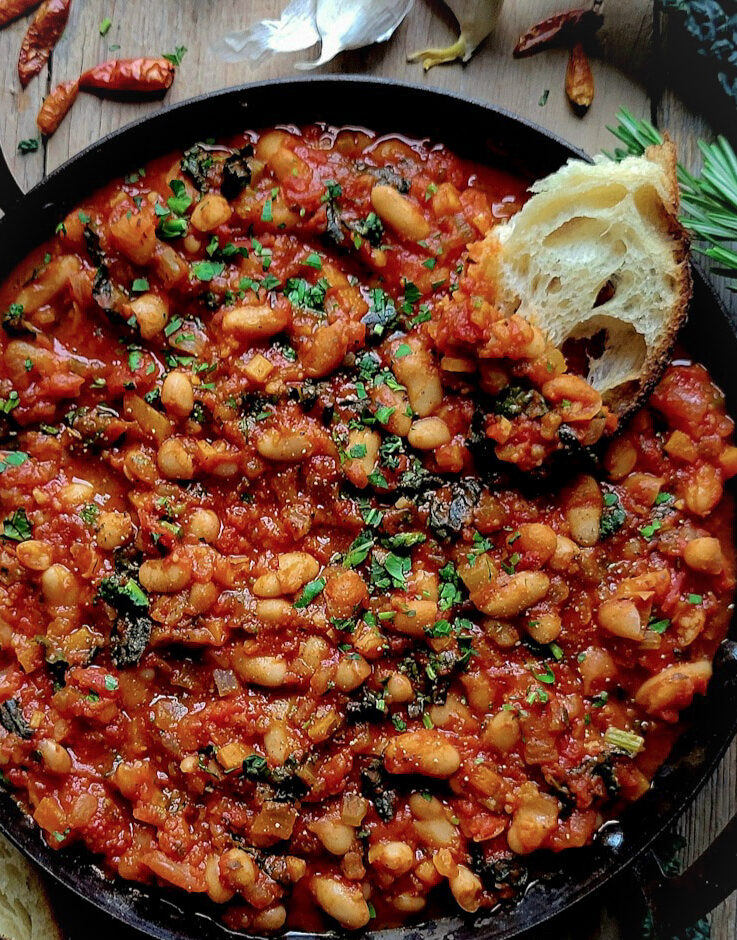 A skillet filled with Tuscan Braised Beans and Kale, with a slice of crusty bread to the side.
