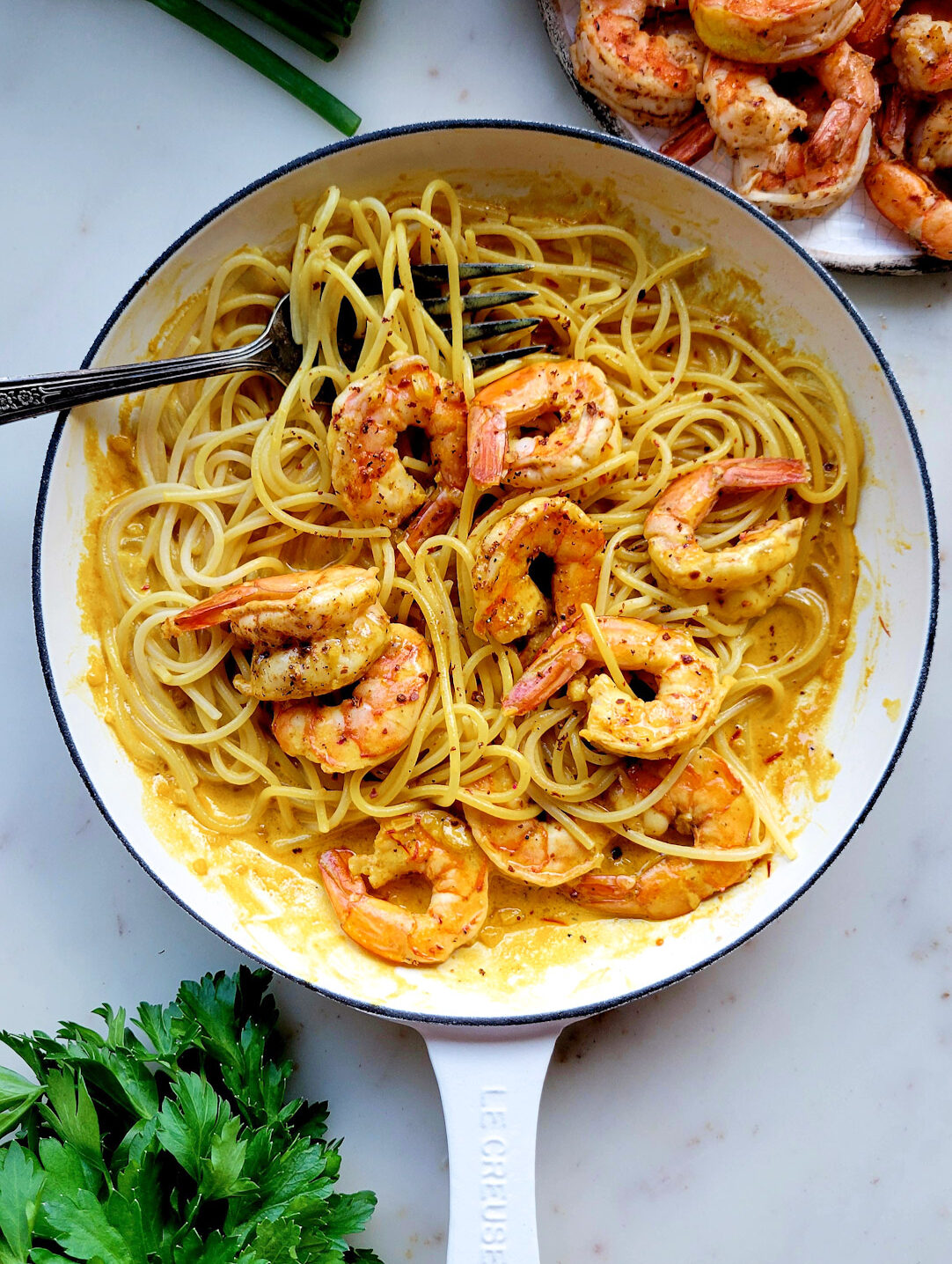 A skillet filled with Harissa Shrimp Pasta with Saffron Sauce, More shrimp and parsley are to the side.