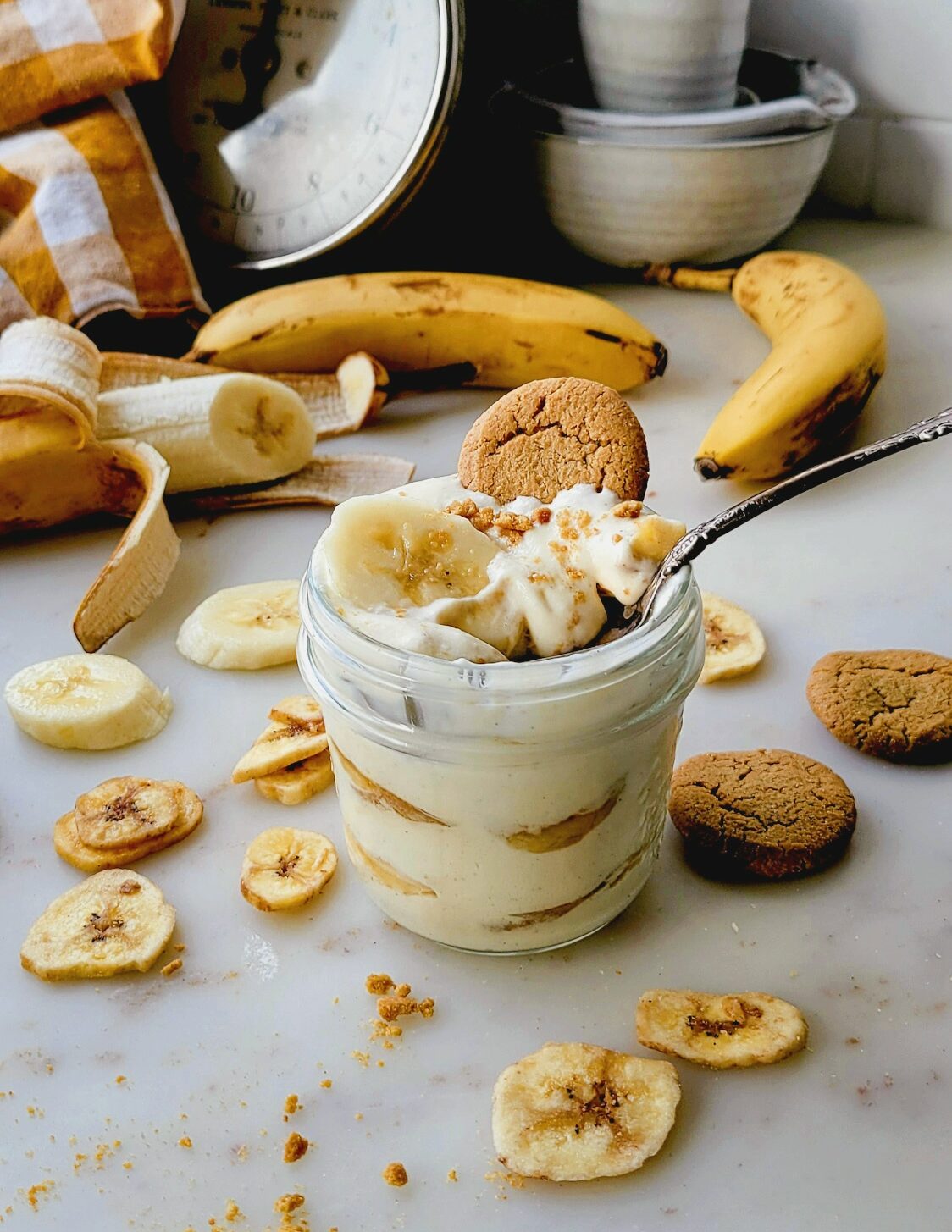 Classic Banana Pudding in a mason jar with a spoon in it. Peeled bananas and ginger snap cookies are in the background.