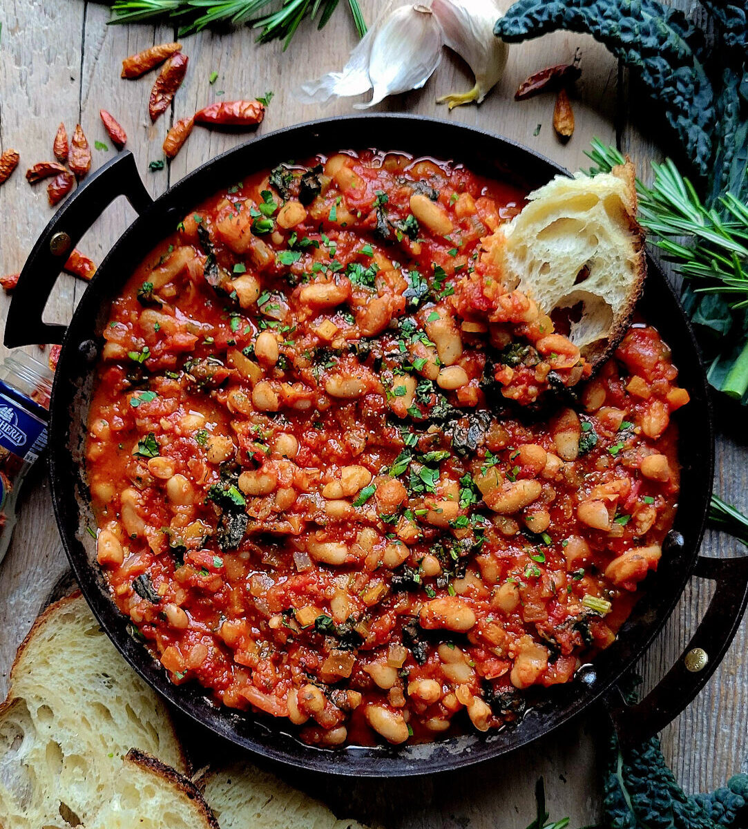 A skillet filled with Tuscan Braised Beans and Kale, with crusty bread to the side, as well as fresh rosemary, garlic cloves and kale leaves.