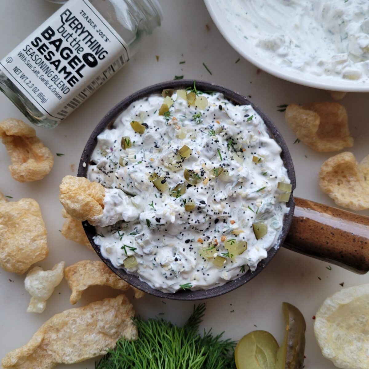A bowl of Dill Pickle Dip with Jalapeno Pepper in on the counter surrounded by chips and Bagel Sesame Seasoning/