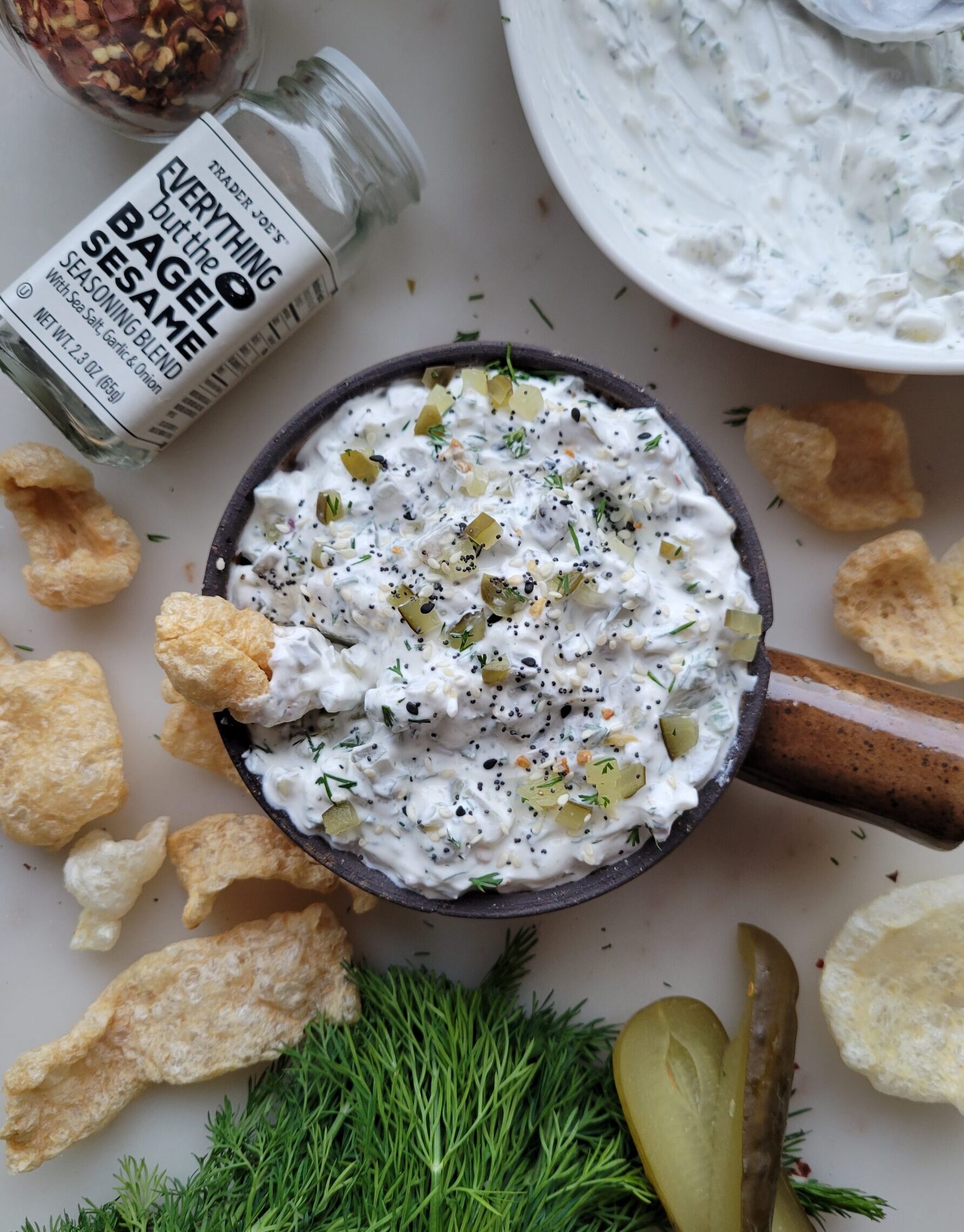 A bowl of Dill Pickle Dip with Jalapeno Pepper in on the counter surrounded by chips and Bagel Sesame Seasoning/