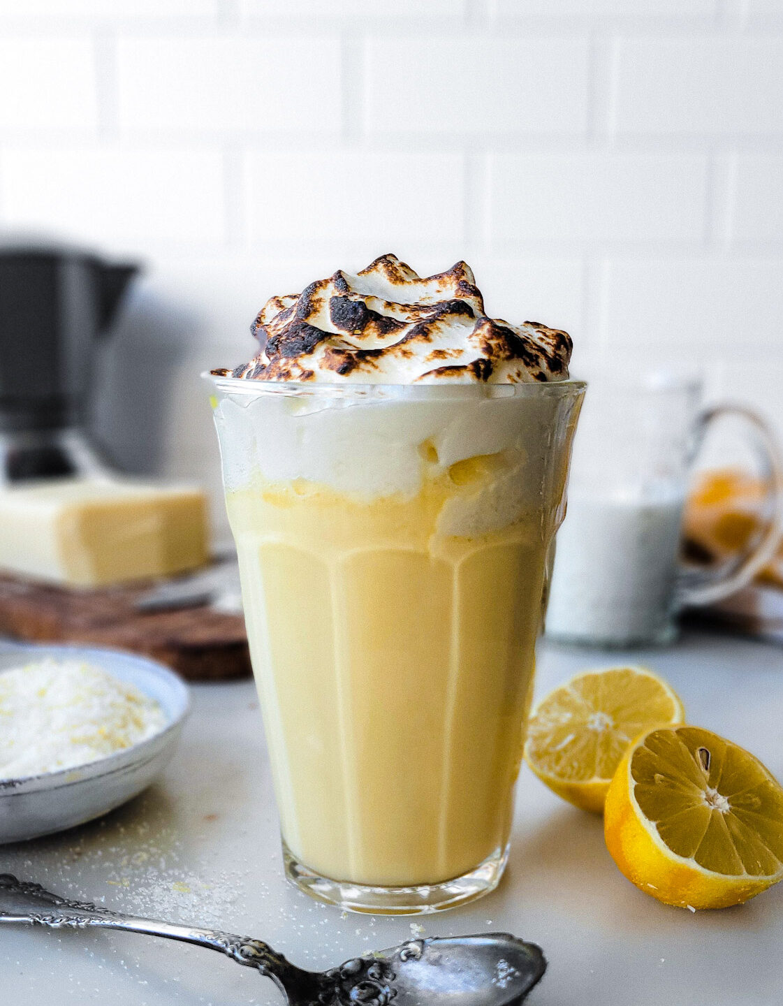 A glass filled with Lemon Meringue Hot Chocolate surrounded by sliced lemons and a bowl of lemon sugar.