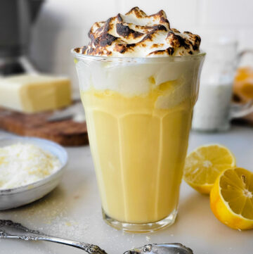 A glass filled with Lemon Meringue Hot Chocolate surrounded by lemons and lemon sugar.