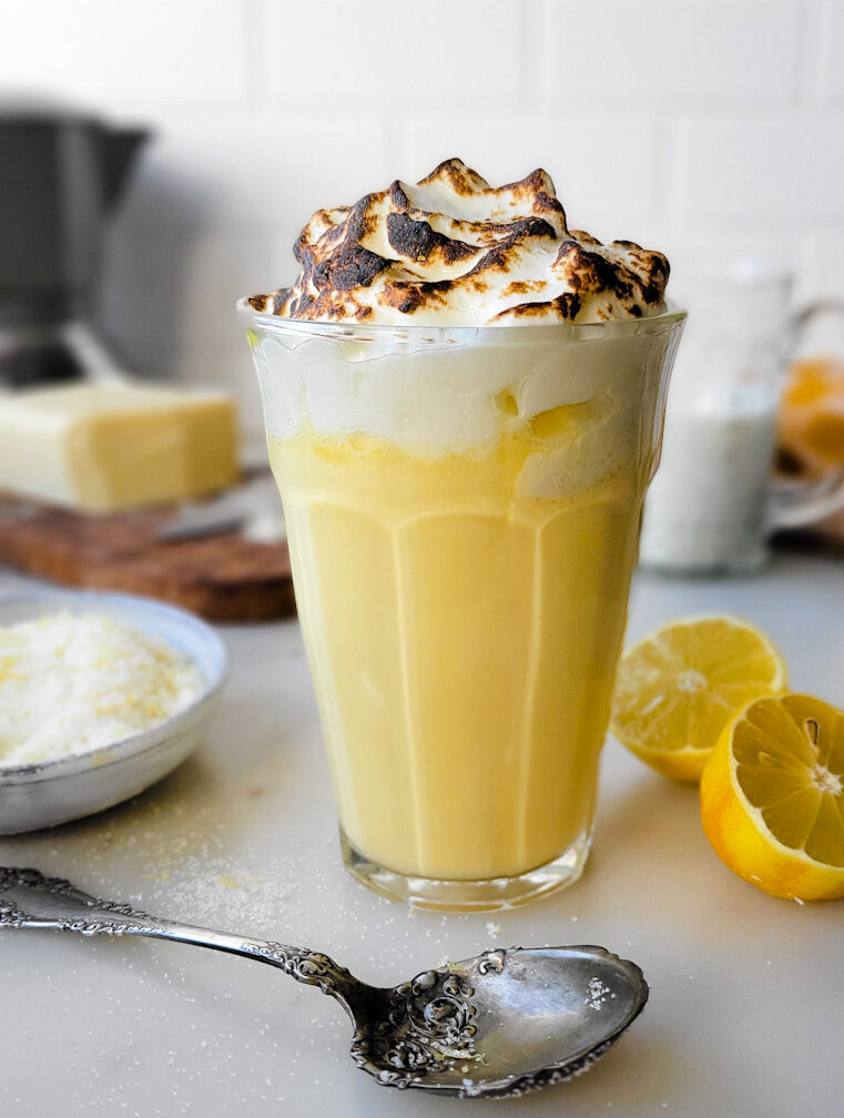 A glass filled with Lemon Meringue Hot Chocolate surrounded by lemons and lemon sugar.