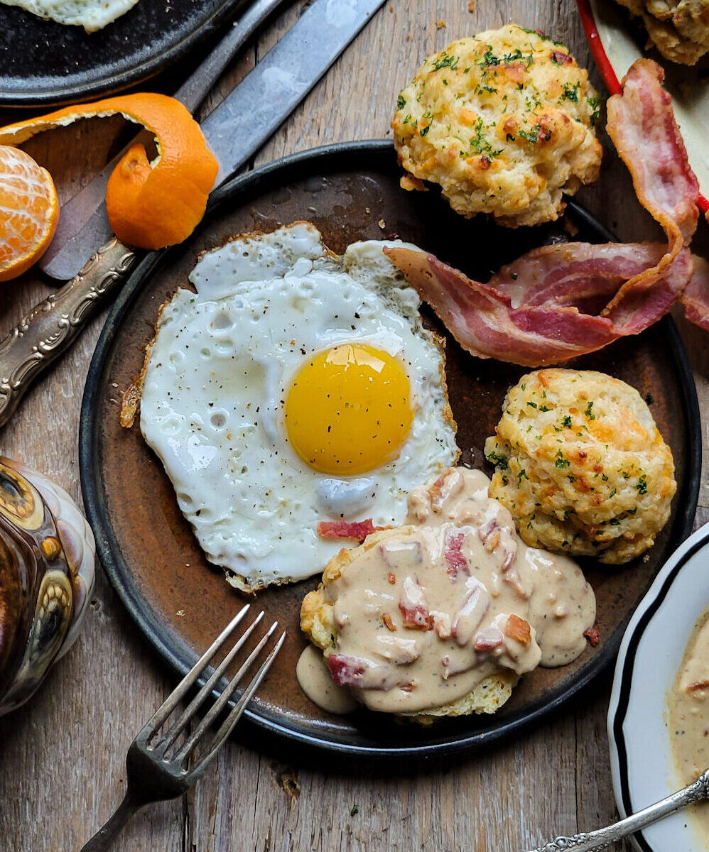 A breakfast plate filled with a sunny side up egg, bacon and an Easy Cheddar Drop Biscuit smothered in Bacon Gravy.