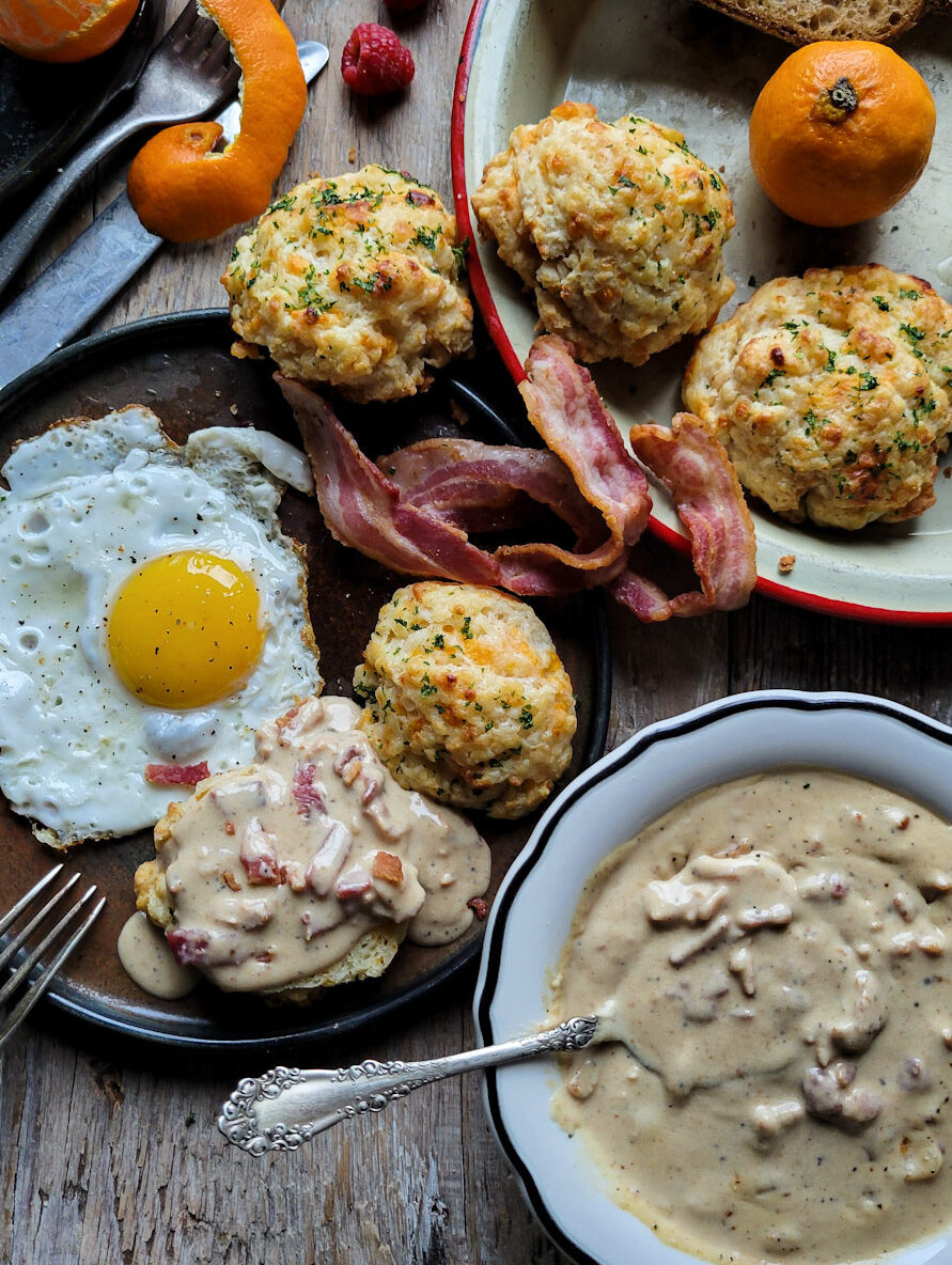 A breakfast plate filled with a sunny side up egg, bacon and an Easy Cheddar Drop Biscuit smothered in Bacon Gravy.