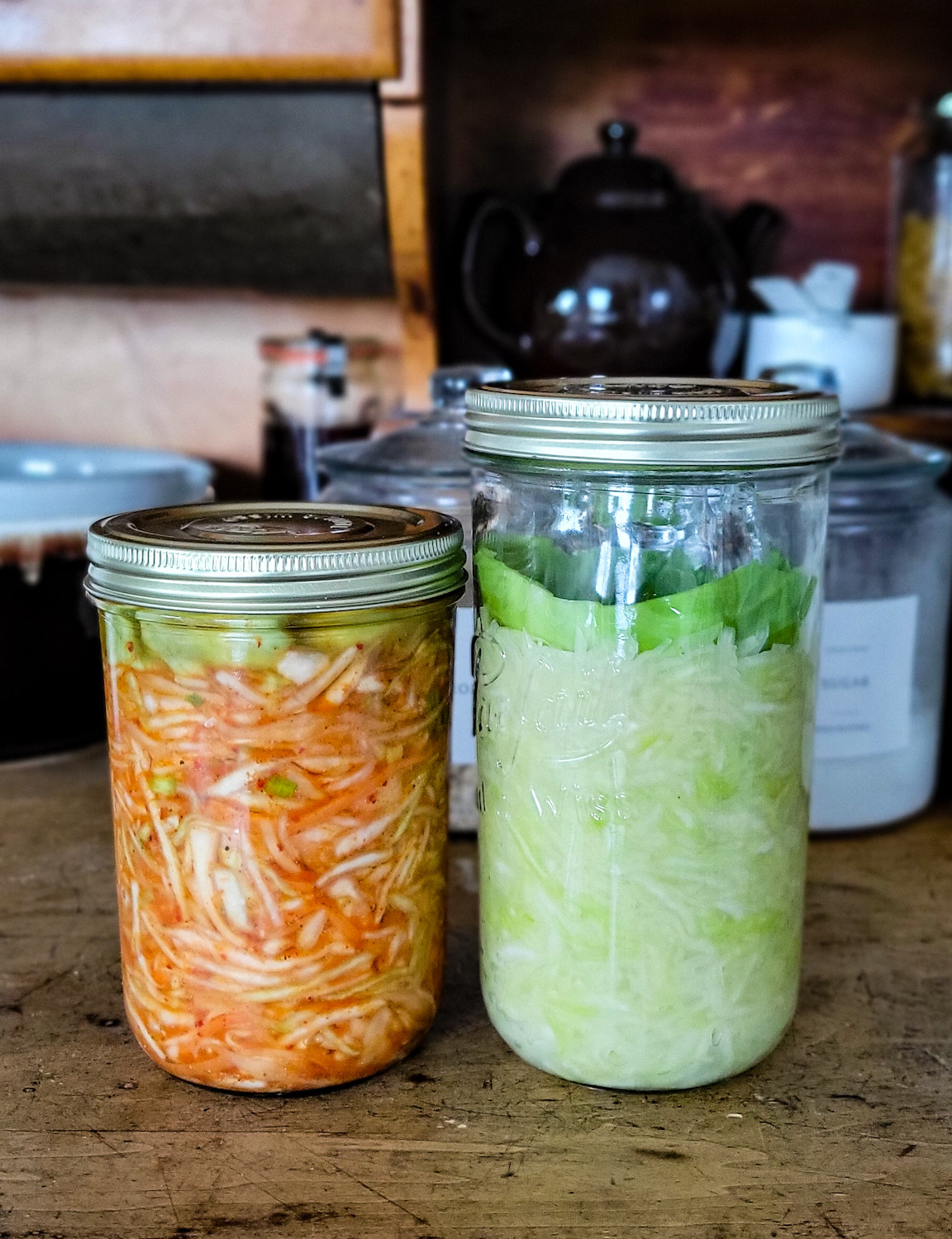A jar of Sauerkraut, and a jar of Kimchi Sauerkraut on the counter, fermenting.