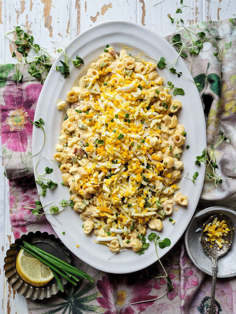 A platter filled with Mimosa Egg Pasta Salad sitting on the table, with spring green shoots and chives to the side.